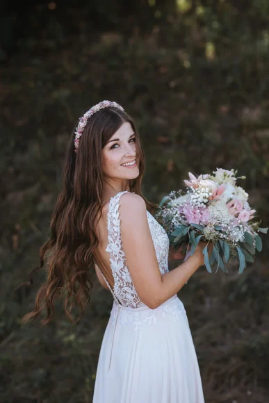 Eine lächelnde Braut mit langem, welligem Haar trägt ein Spitzenbrautkleid mit offenem Rücken, ein rosa Blumenstirnband und hält im Freien einen Strauß pastellfarbener Blumen.
